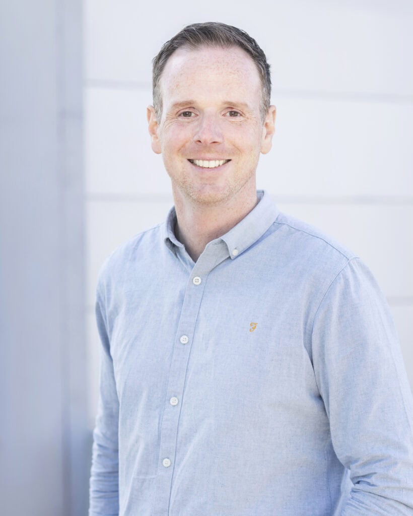 Headshot of a smiling man with short brown hair wearing a light blue button-up shirt standing outside against a light-colored background.