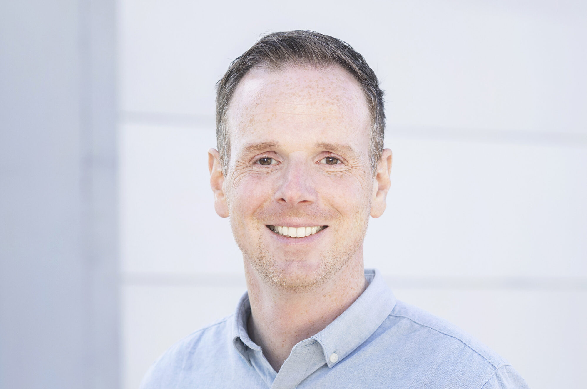 Headshot of a smiling man with short brown hair wearing a light blue button-up shirt standing outside against a light-colored background.