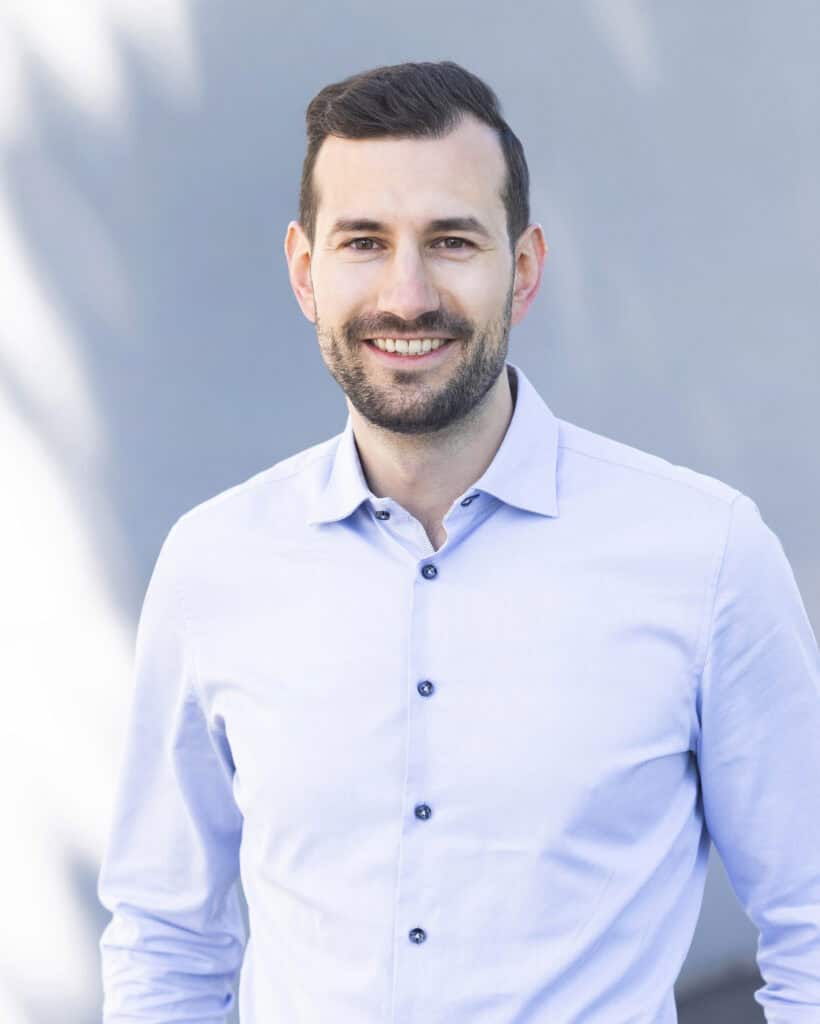Handsome young man with short dark hair and a beard, wearing a light blue dress shirt, smiling confidently outdoors against a blurred modern building background.