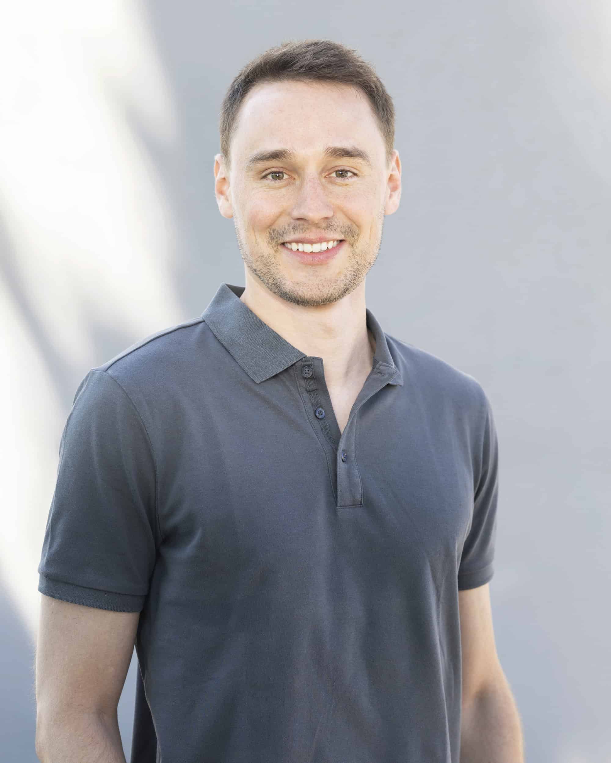 Smiling young man with short brown hair and a light beard, wearing a dark grey polo shirt, standing outdoors against a blurred background with natural light.