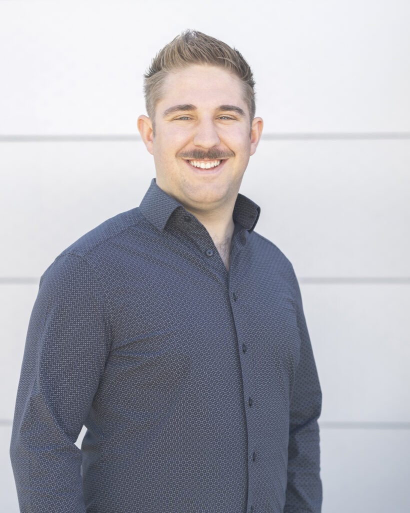 Smiling man with short blond hair and a mustache wearing a dark blue, patterned dress shirt standing against a white wall.