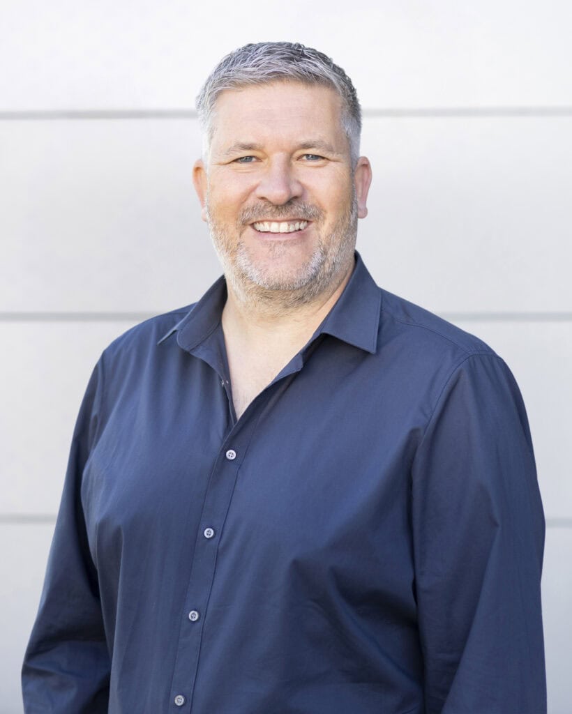 Smiling middle-aged man with gray hair and beard wearing a navy blue shirt outdoors against a light-colored wall.