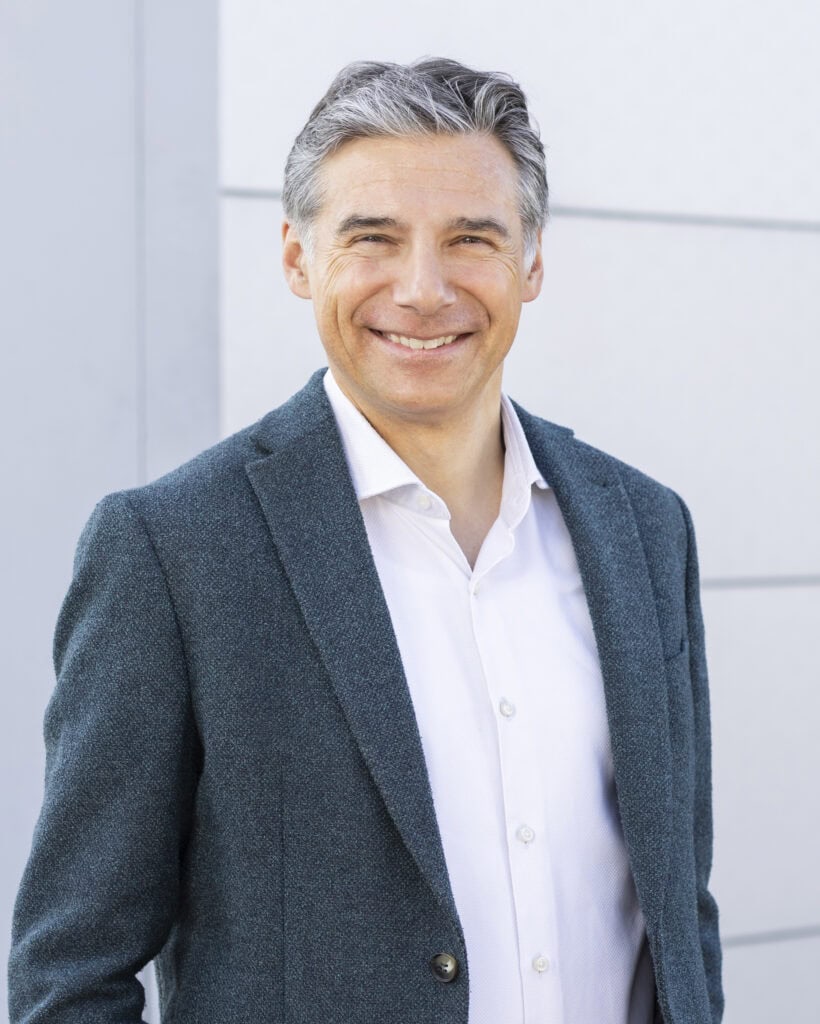 Senior man in a white shirt and dark blazer smiling outdoors.