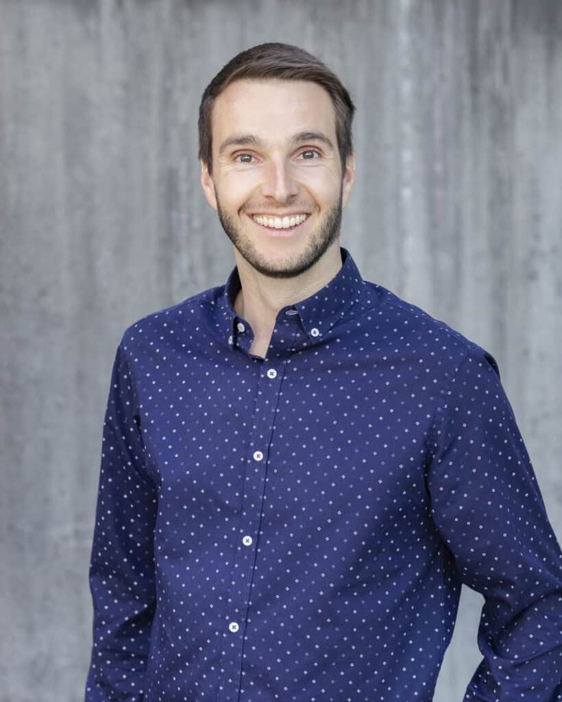 Smiling young man with a beard wearing a navy blue polka dot shirt against a concrete wall background.