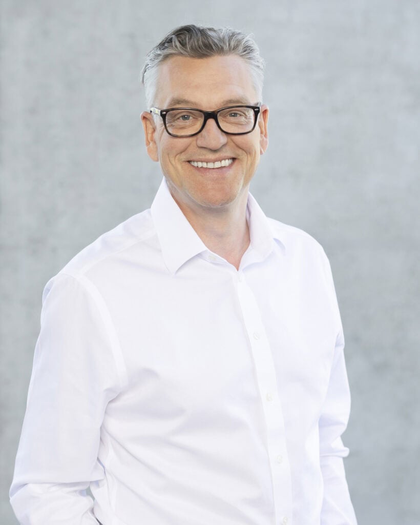 Professional middle-aged man wearing glasses and a white dress shirt, smiling confidently against a neutral gray background.