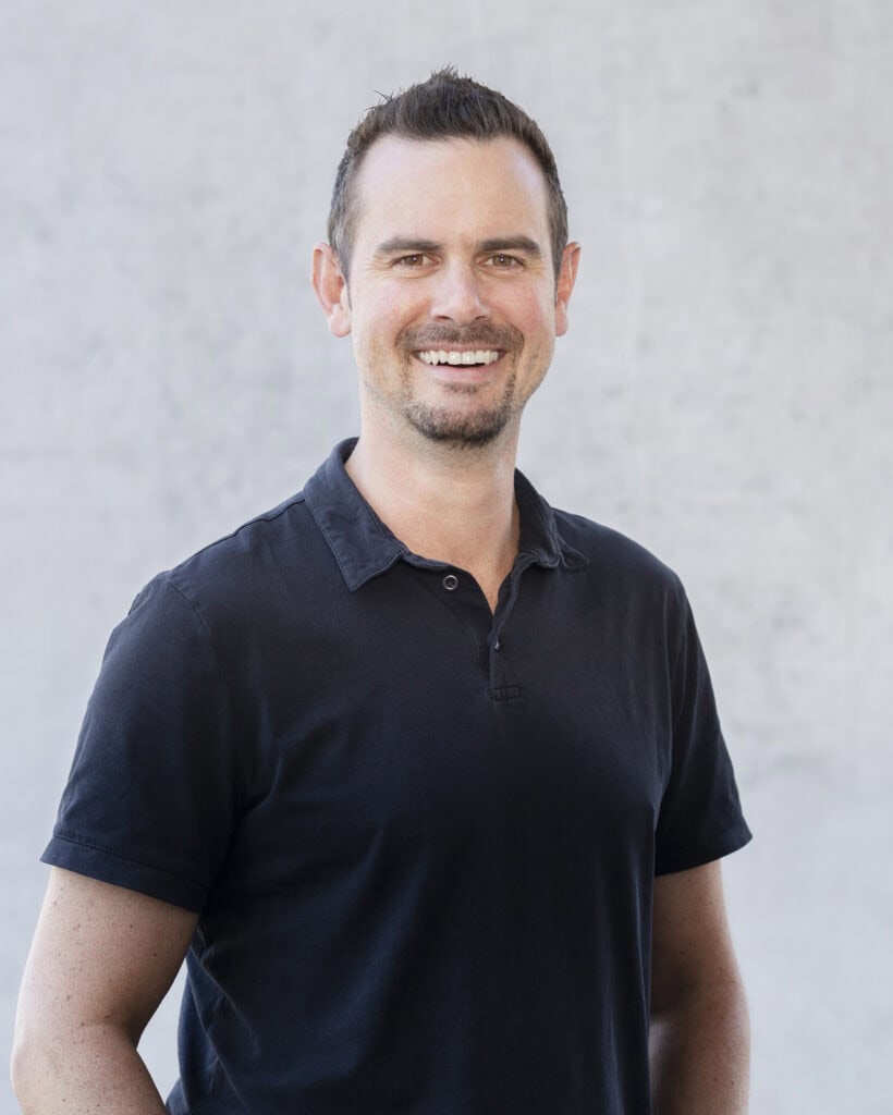 Friendly smiling man with short dark hair, neatly trimmed beard, wearing a casual black polo shirt, standing outdoors against a plain light gray background.