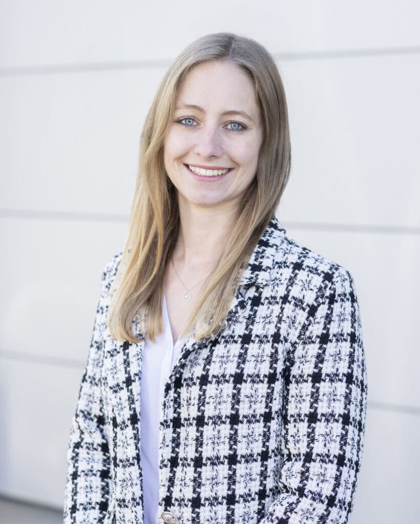 Bright smiling woman with blue eyes and blonde hair, wearing a black and white checkered blazer and jewelry, standing outdoors against a light-colored wall.
