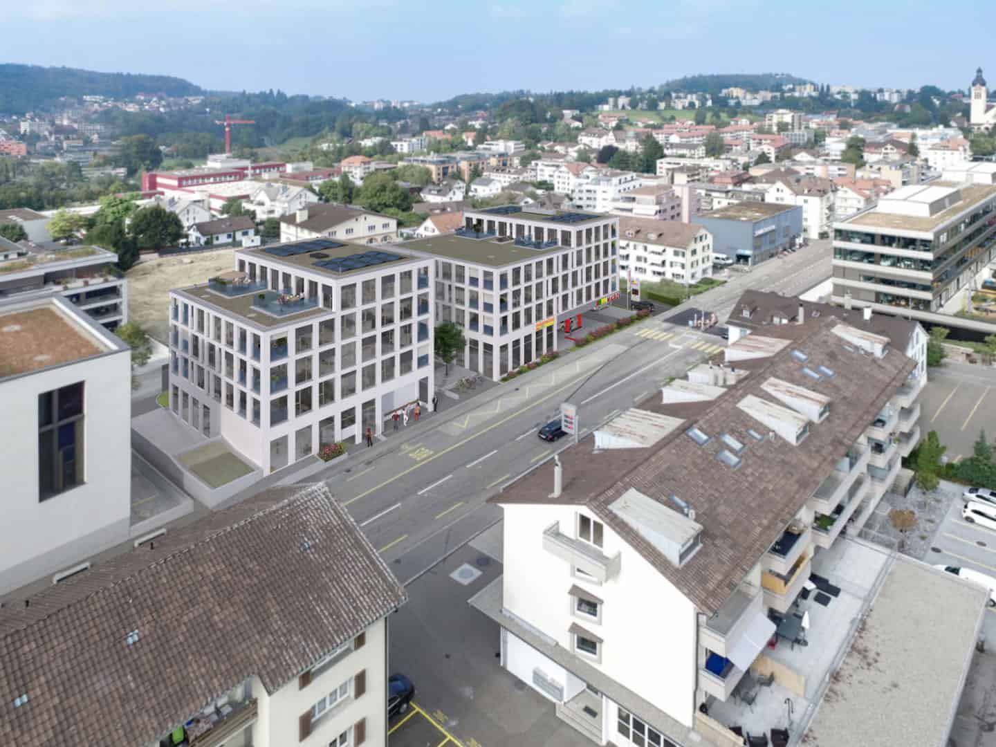 Contemporary multi-story residential and commercial buildings in an urban neighborhood, featuring modern architecture, rooftop terraces, and solar panels, with traditional houses and a hilly landscape in the background.