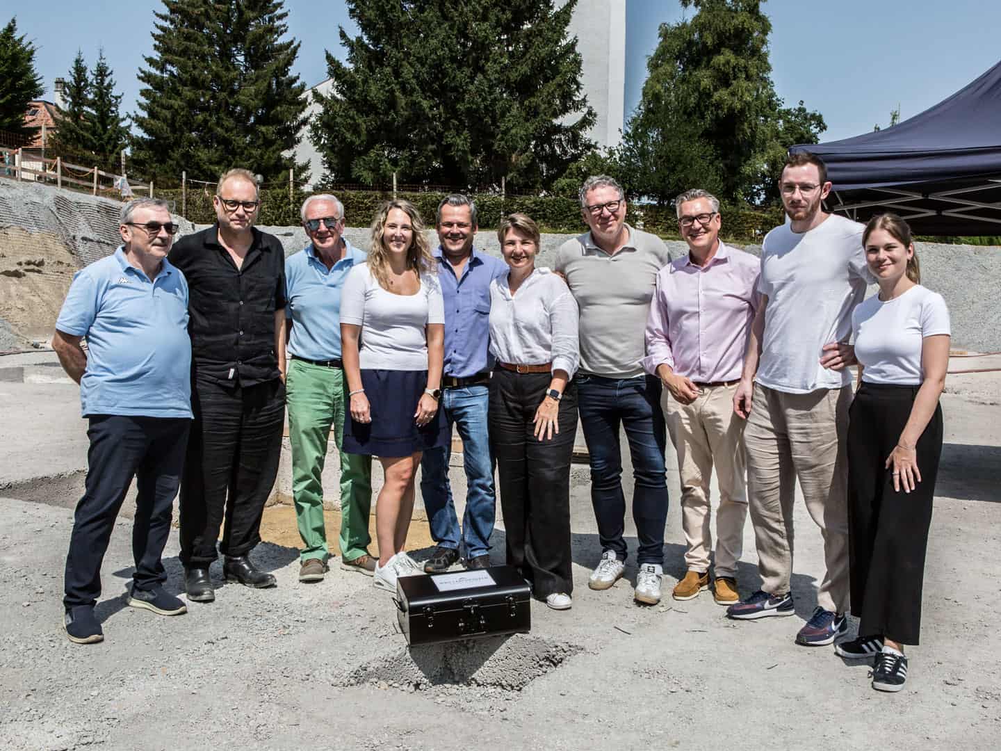A diverse group of professionals standing outdoors in a construction site, smiling for the photo under a sunny sky, with trees and construction materials in the background.