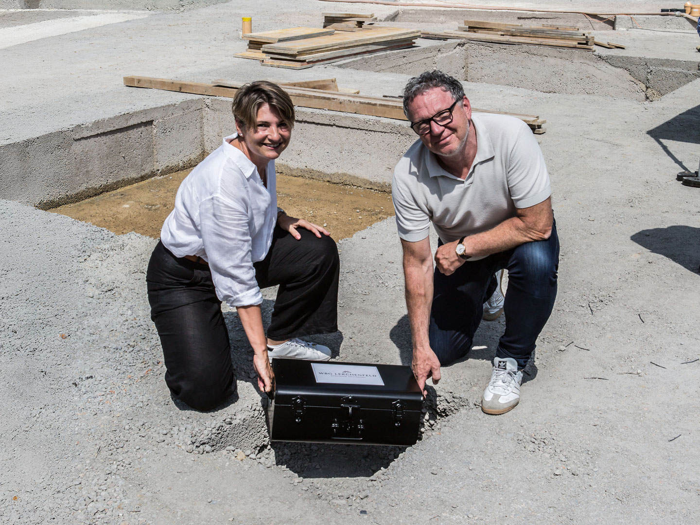 A woman and a man kneel next to a construction foundation at a building site, smiling and holding a black toolbox. They appear to be working on construction or renovation project outdoors on a sunny day.