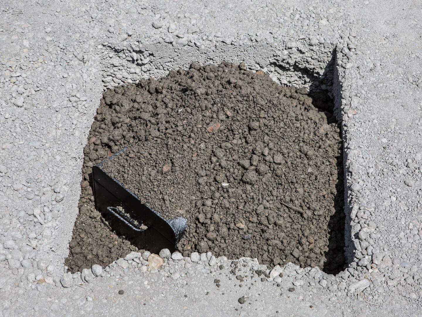 Layer of loose gravel and soil with a small black container partially buried in the ground. The surrounding surface appears to be composed of compacted gravel and dirt, possibly a construction site or garden bed.